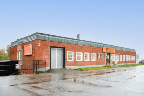 En envånings industribyggnad i rött tegel med många fönster, ett platt tak och en mulen himmel i bakgrunden. Marken utanför ser ut att vara våt av regn.