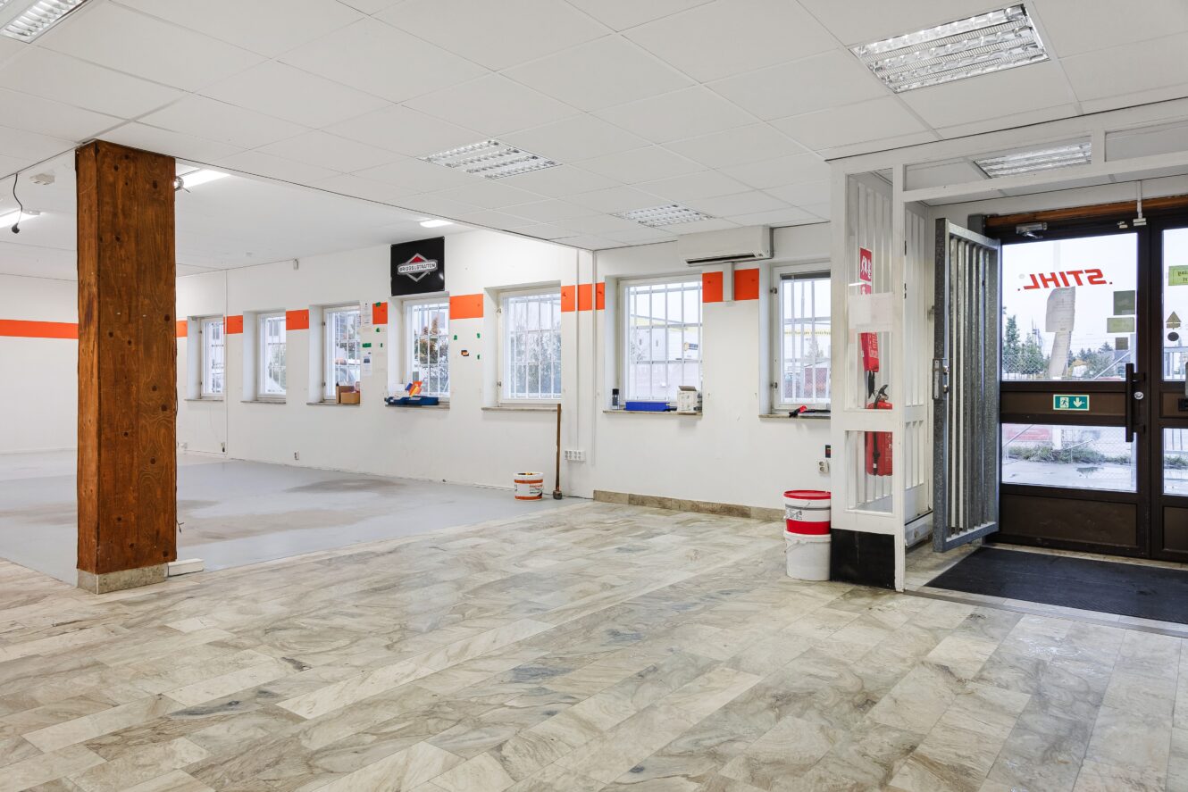 Tom affärslokal med klinkergolv, vita väggar, träpelare, väggmonterade värmare och öppen entrédörr; flera fönster med spröjs och orange accenter.