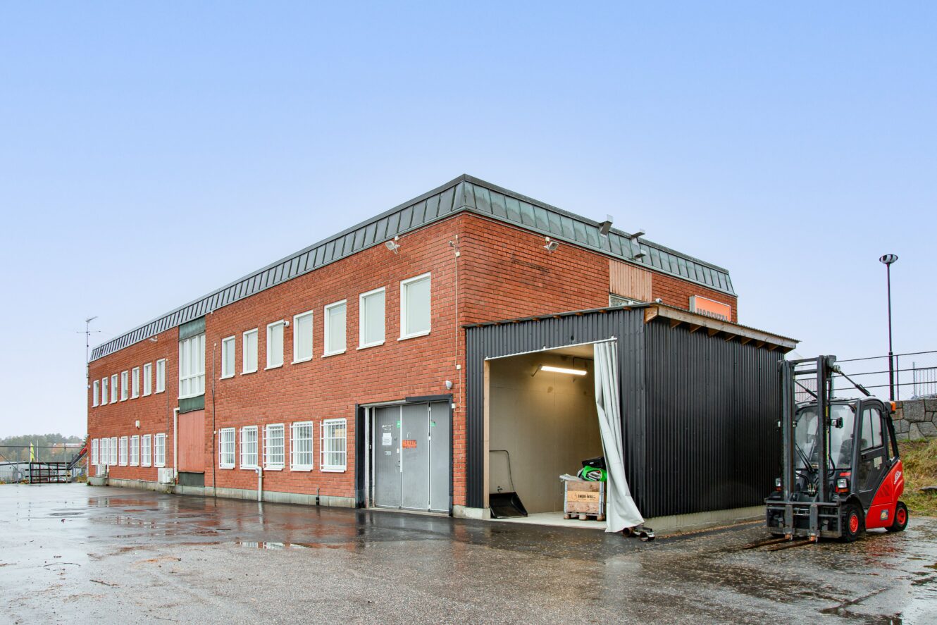 En tvåvånings industribyggnad i rött tegel med många fönster, en grå metallentré och en gaffeltruck som står parkerad nära ett öppet garage på en våt betongtomt.
