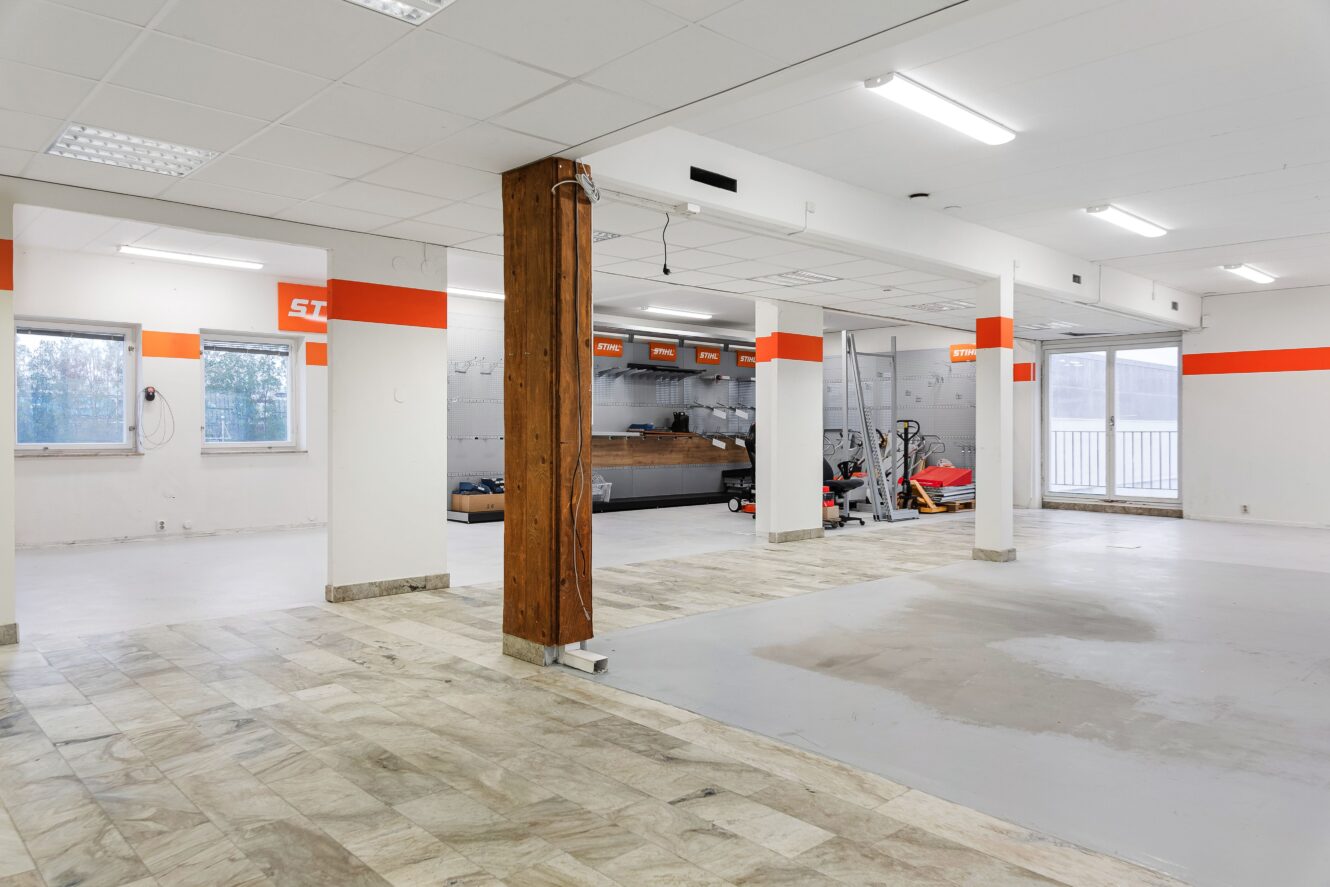 Stort, rent och väl upplyst garage eller verkstadslokal med klinkergolv, vita väggar, träpelare och träningsutrustning i det bakre hörnet.