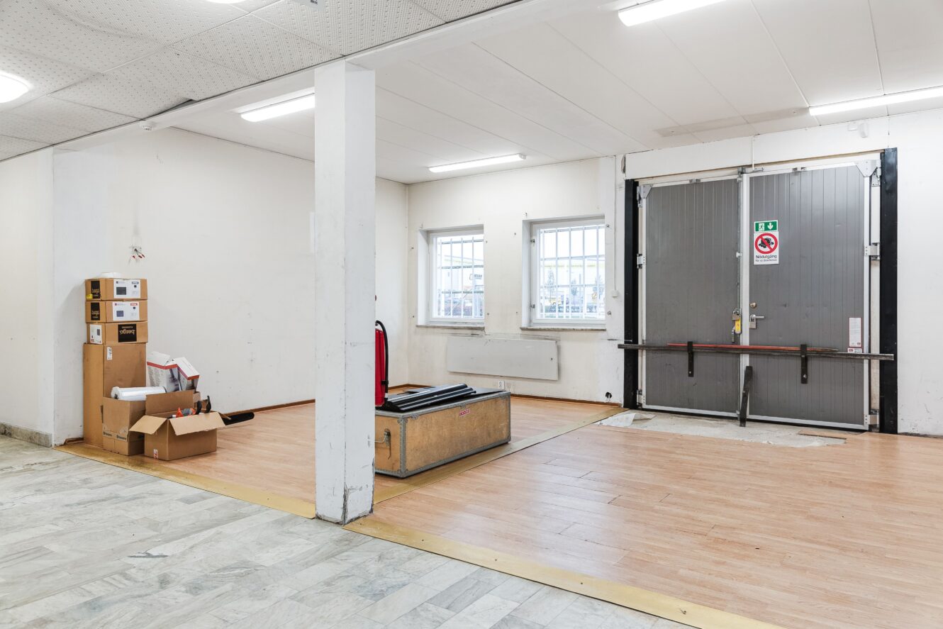 En i stort sett tom industrilokal med trämönstrat golv, en lastbryggedörr i metall, gallerförsedda fönster, en brandsläckare och några utspridda lådor och föremål i ett hörn.