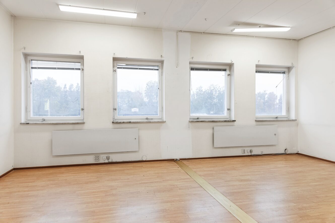 Ett tomt rum med fyra rektangulära fönster, vita väggar, taklampor, trägolv och synliga radiatorer under fönstren.