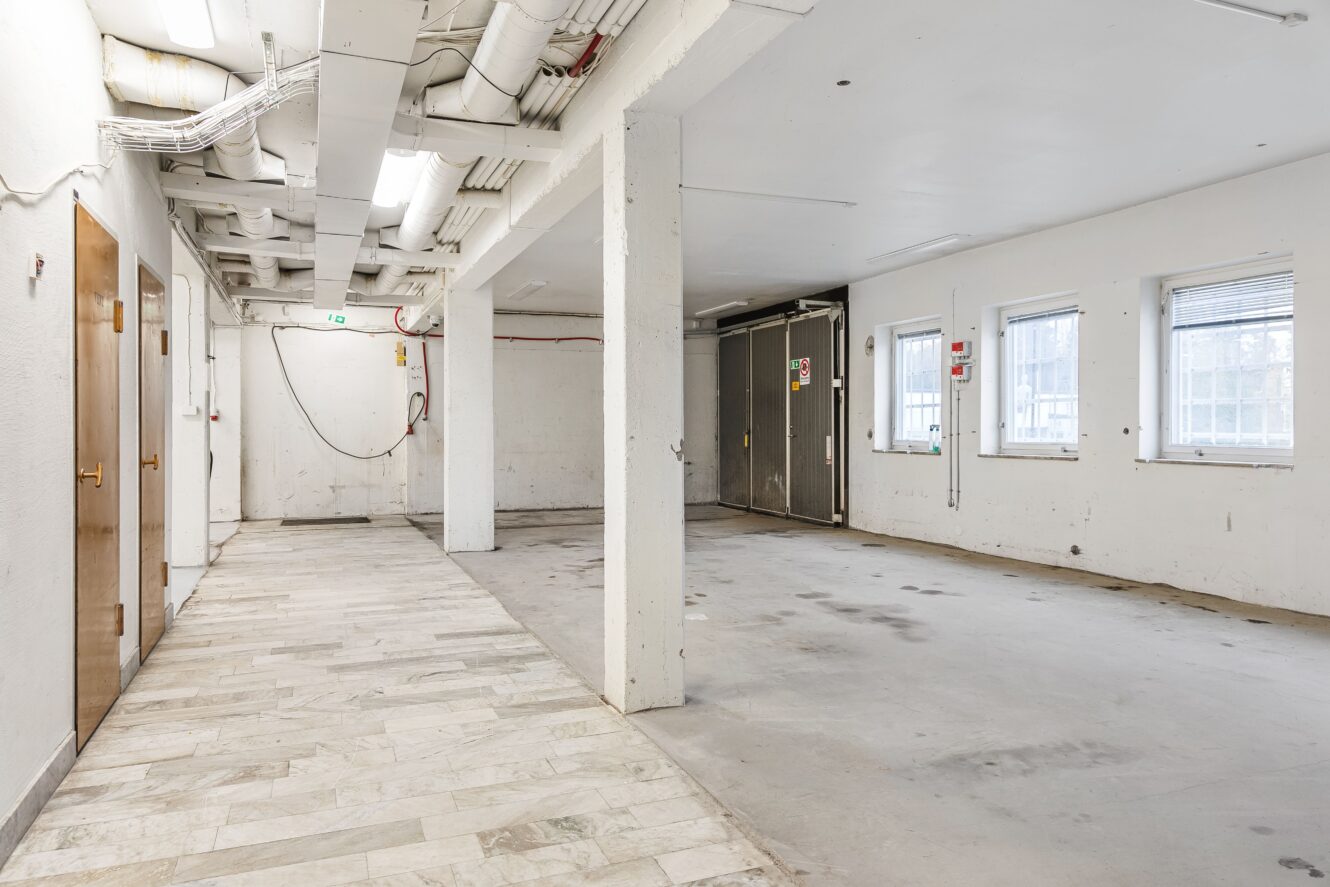 Tomt industrirum med synliga rör i taket, vita väggar, klinker- och betonggolv, flera fönster och lysrörsbelysning.