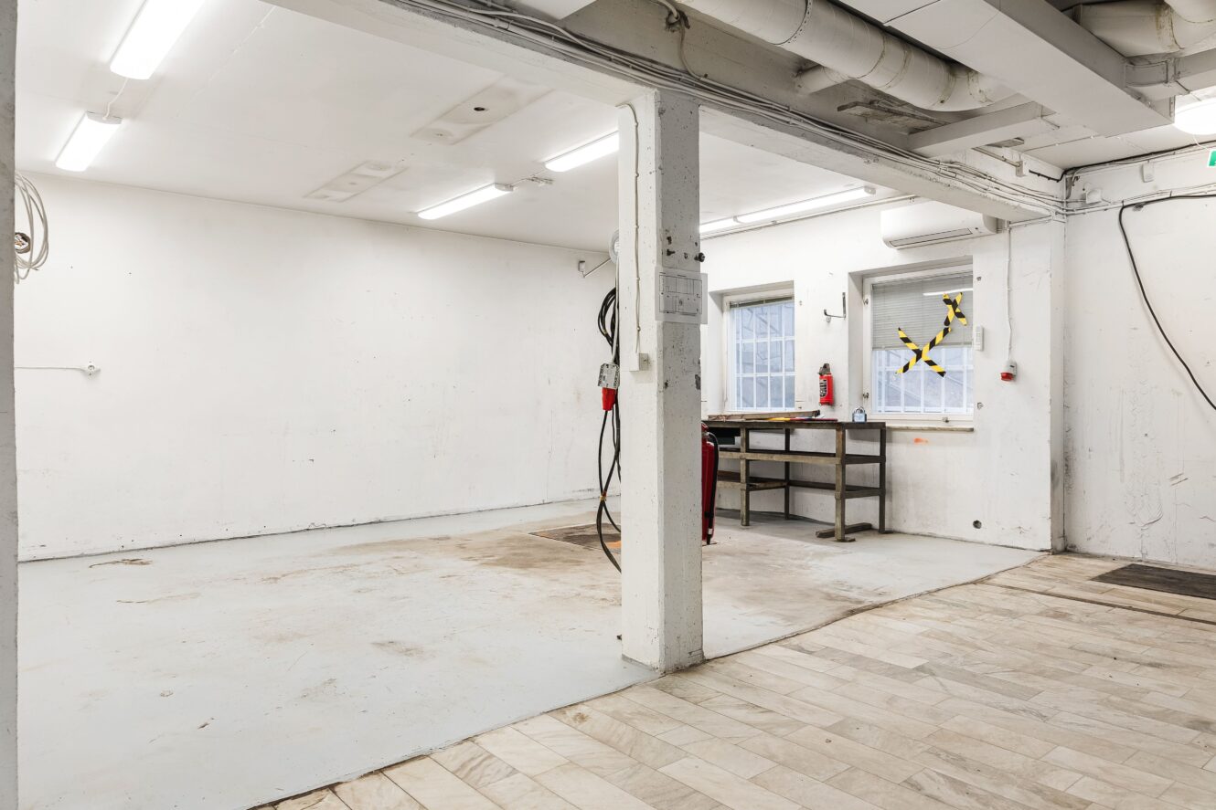 Tom industriverkstad med vita väggar, synliga rör, lysrör, en arbetsbänk under två fönster samt klinker- och betonggolv.