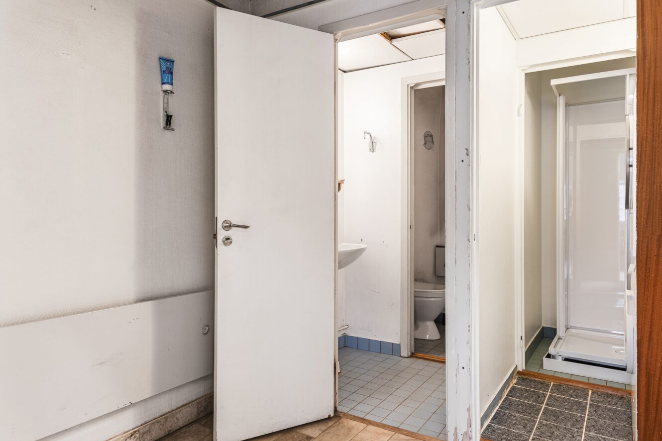 Ett litet badrum med en vit dörr som är delvis öppen och avslöjar ett handfat, en toalett och ett klinkergolv; en duschkabin syns i närheten utanför badrummet.