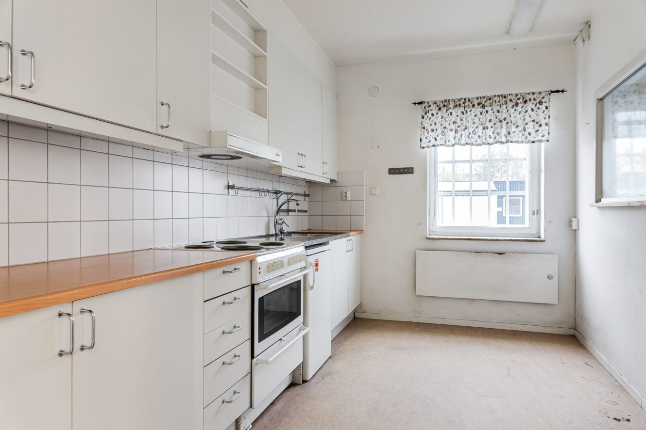 Ett litet, vitt kök med kaklat stänkskydd, elspis, ugn, skåp och ett fönster med en blommig gardin som släpper in naturligt ljus.