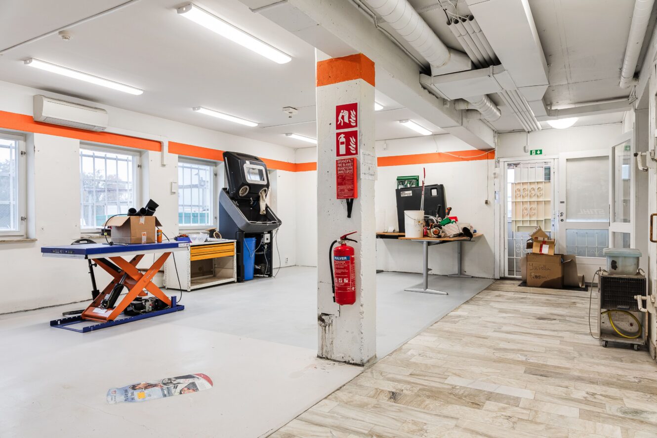 En rymlig, väl upplyst verkstad med utspridda verktyg, arbetsbänkar, två arkadmaskiner och en brandsläckare monterad på en pelare.