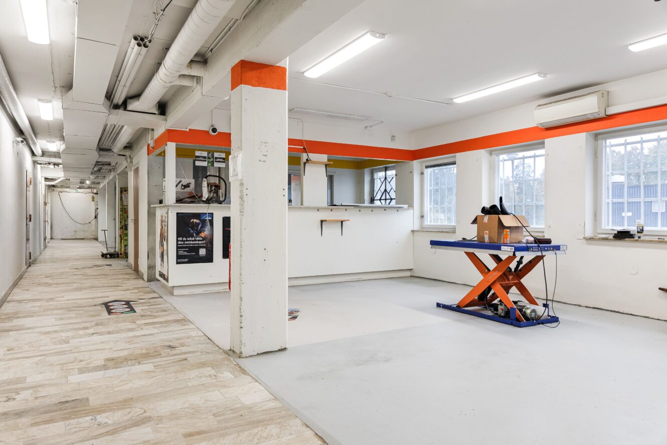 Ett rymligt rum i industriell stil med vita väggar, orangefärgade detaljer, stora fönster, synliga rör och ett blått saxlyftbord som håller i en pappkartong.