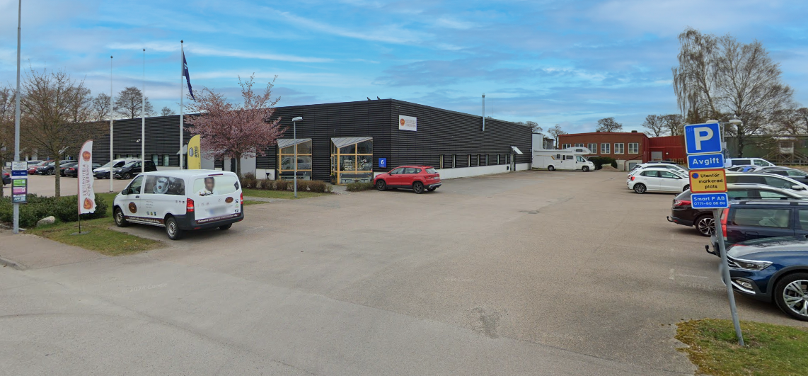 En parkeringsplats med flera bilar utanför en modern envåningsbyggnad, flankerad av träd och skyltar som anger avgiftsbelagd parkering. Blå himmel med spridda moln ovanför.