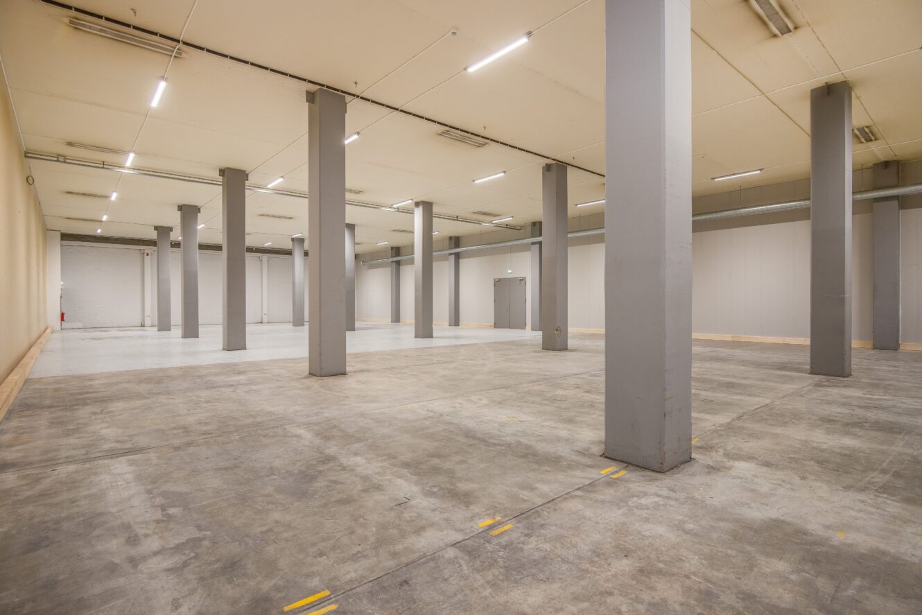Stor tom lagerlokal med betonggolv, höga grå stödpelare och stark takbelysning.