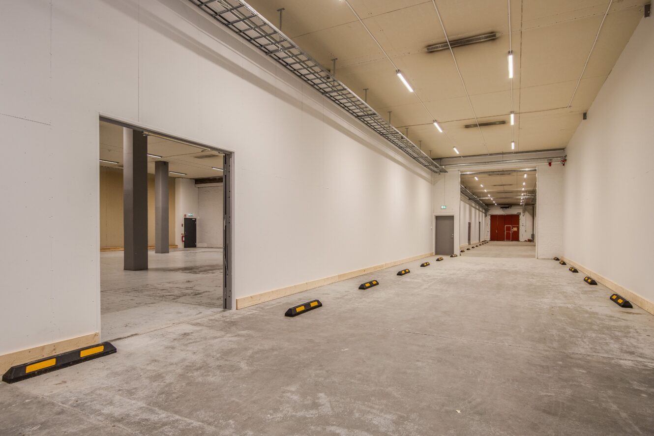 En bred, tom inomhuskorridor med vita väggar, takbelysning och svartgula parkeringshållplatser längs ena sidan, som leder till en öppen dörröppning och en röd dörr i fjärran.