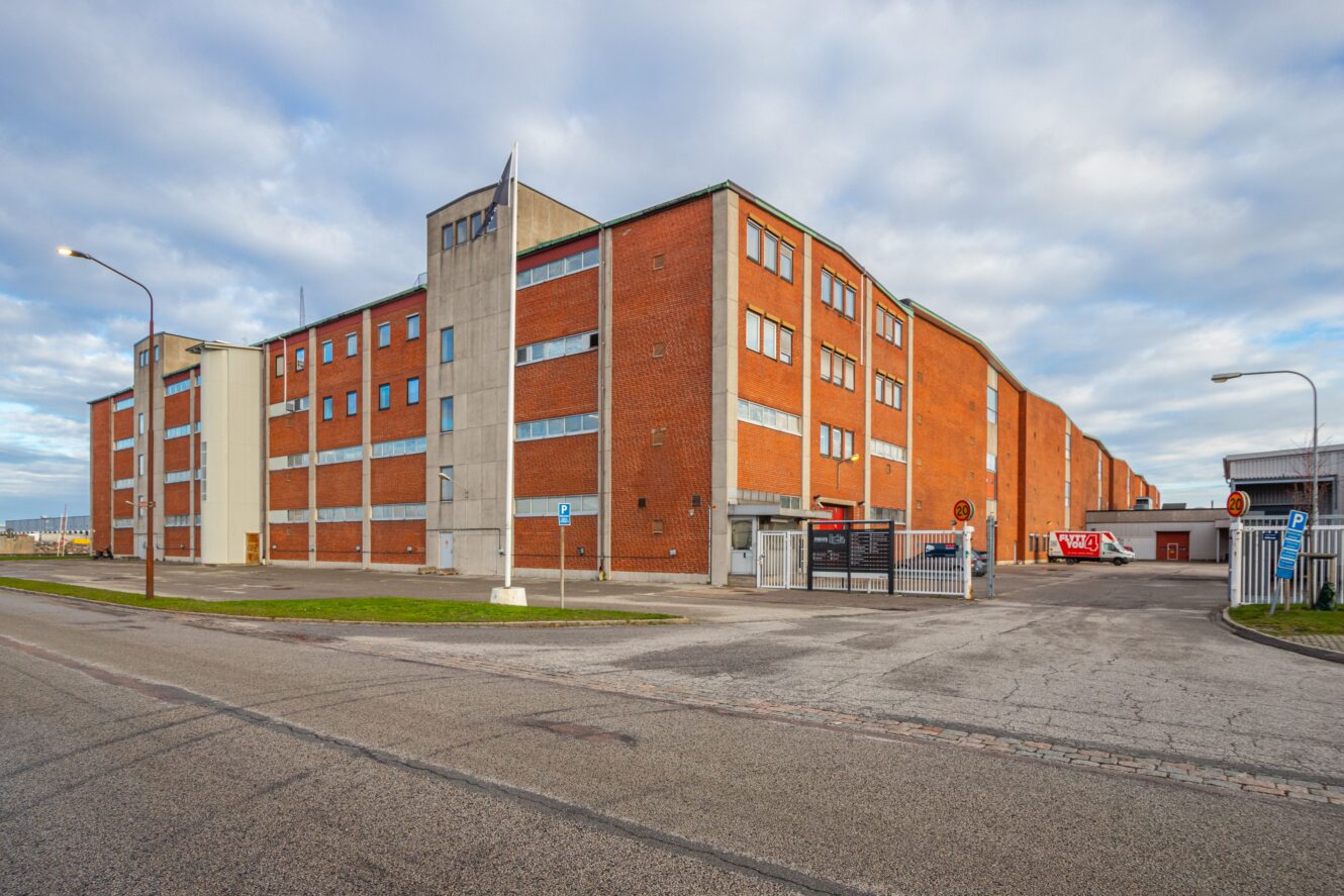 Stor industriell lagerbyggnad i rött tegel med flera fönster, en grindförsedd entré och en i stort sett tom omgivande gata under en molnig himmel.
