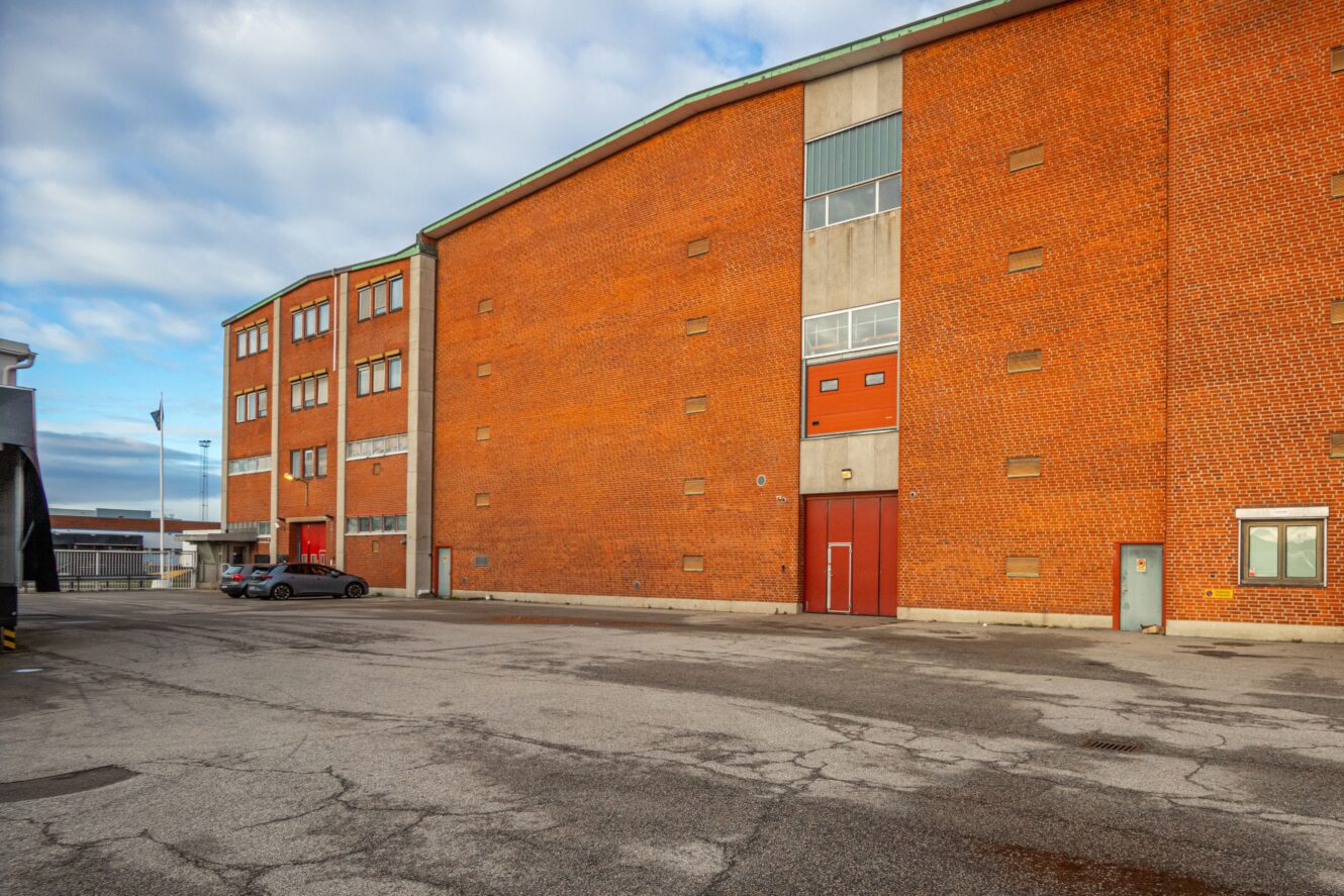 Stor industribyggnad i rött tegel med flera fönster och dörrar, belägen intill en tom asfalterad tomt under en delvis molnig himmel.
