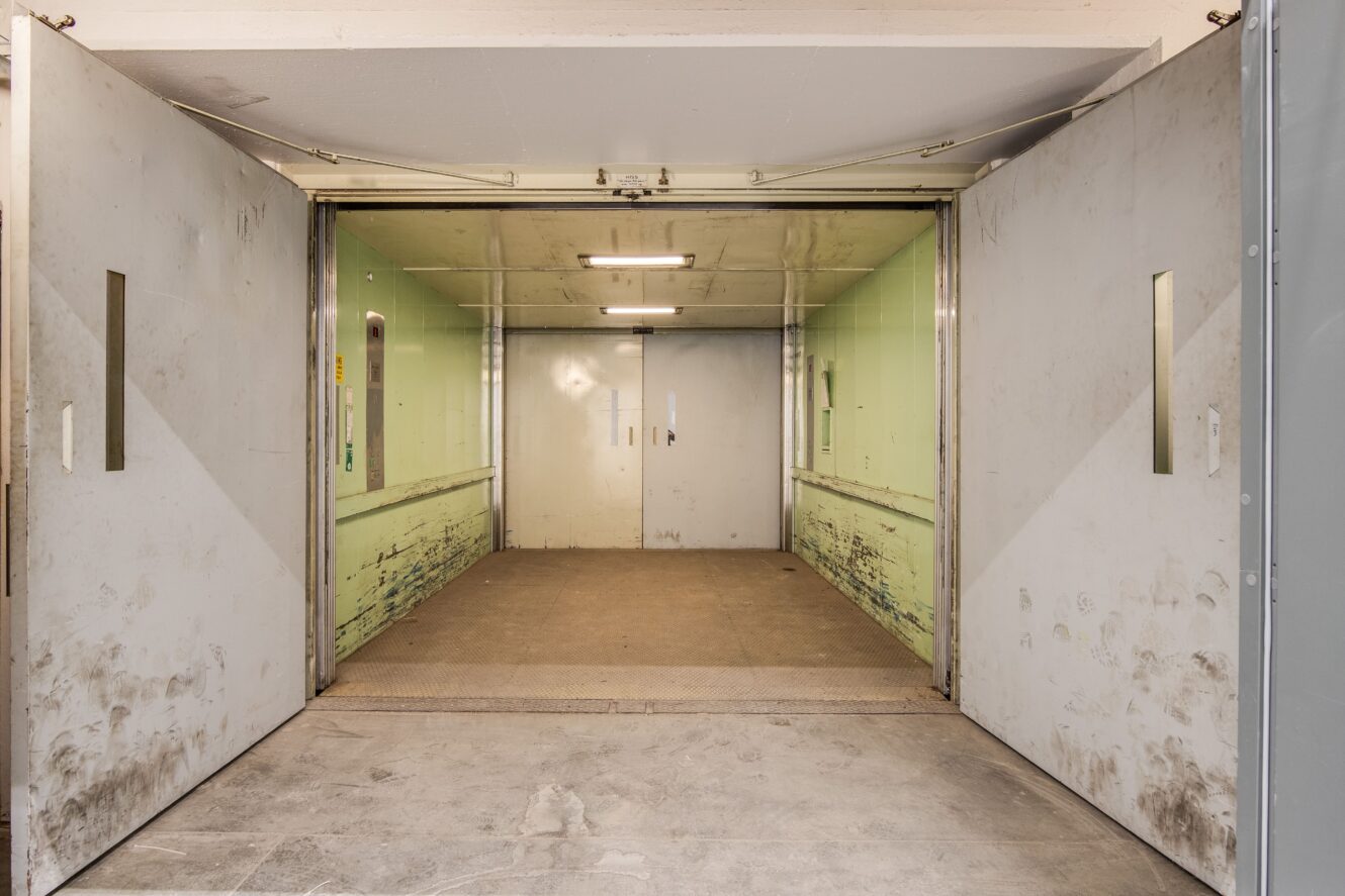 Industriell godshiss med öppna metalldörrar, slitna gröna väggar, sliten inredning och lysrörsbelysning.