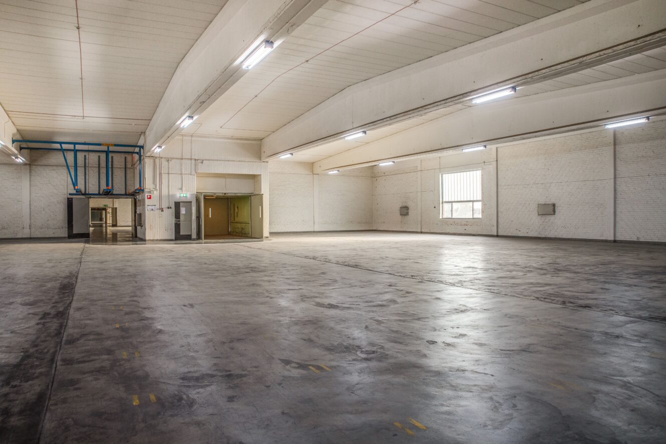 Stor, tom lagerlokal med betonggolv, vita tegelväggar, högt i tak, lysrörsbelysning och ett fönster som släpper in naturligt ljus.