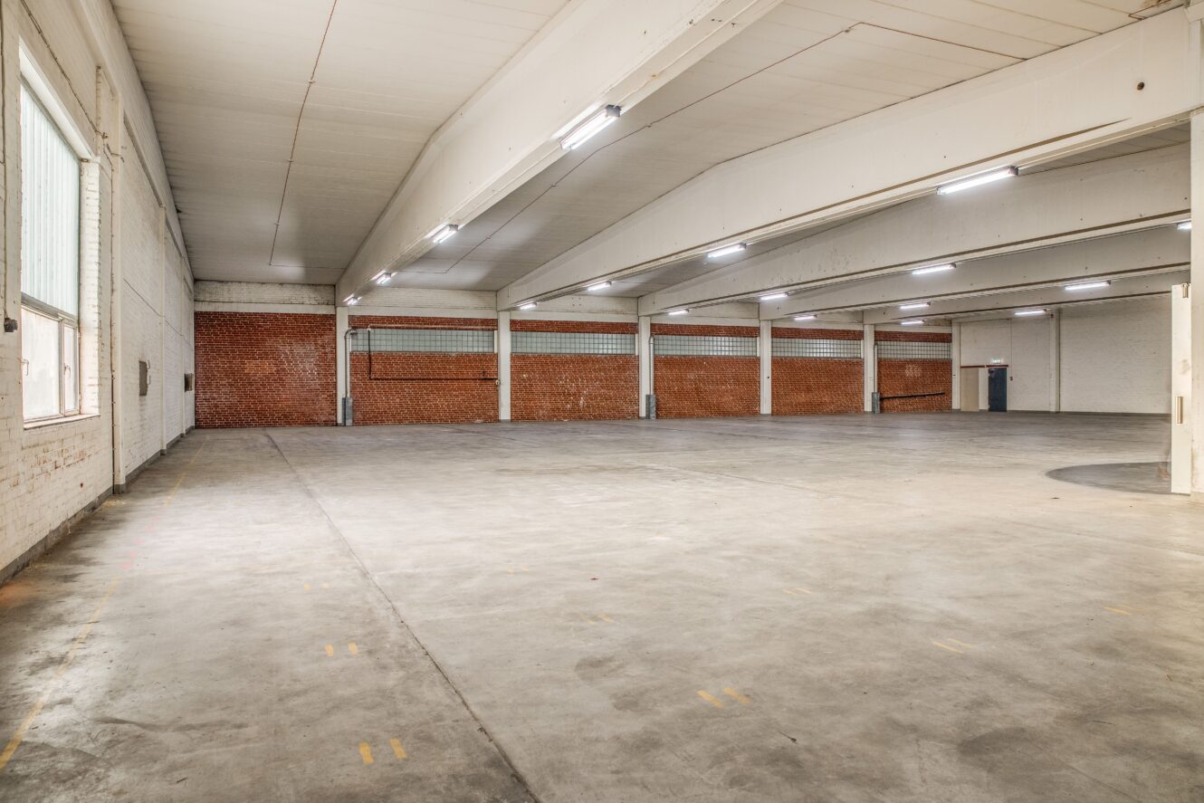 Rymlig, tom lagerlokal med betonggolv, tegelväggar och vita väggar, lysrör i taket och stora fönster till vänster.