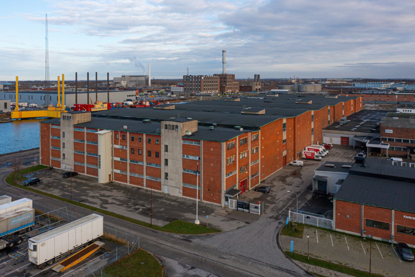 Ett stort industriellt lagerkomplex i rött tegel ligger nära vattnet, omgivet av parkeringsplatser, lastbilar och intilliggande industribyggnader under en molnig himmel.