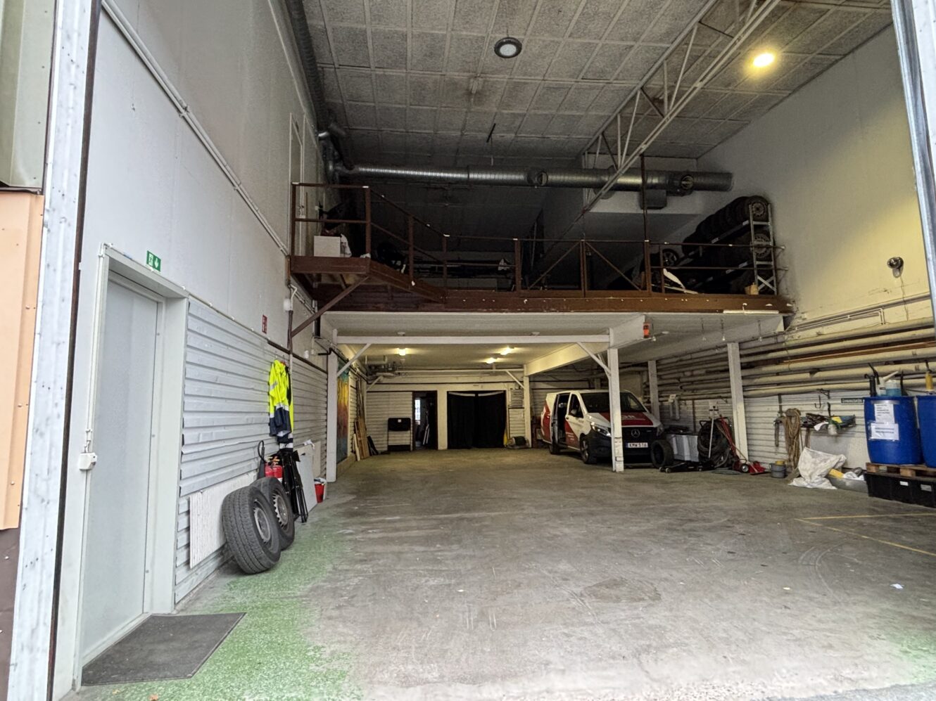 Garage inomhus med entresolplan, parkerade fordon, däckstapel till vänster, hyllor och industriutrustning längs den högra väggen.
