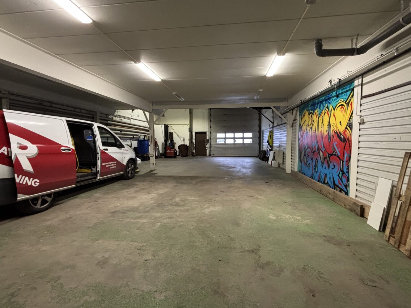 Rymligt garage med en delvis öppen rödvit skåpbil, färgglad graffiti på den högra väggen, verktyg och utrustning längs bak- och sidoväggarna.