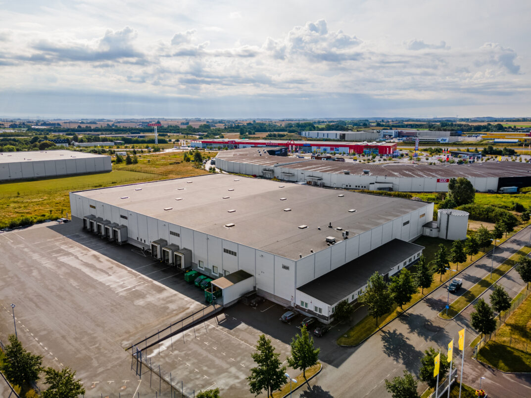 Ett stort industrilager med lastkajer och lastbilar parkerade utanför, omgivet av tillfartsvägar och andra kommersiella byggnader i en företagspark.