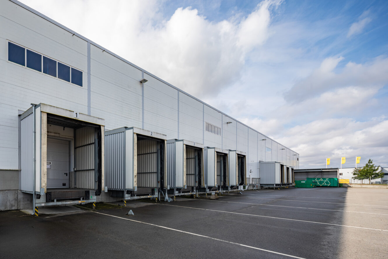 En rad lastkajer med stängda dörrar vid en lagerlokal, bredvid en tom parkeringsplats under en delvis molnig himmel.