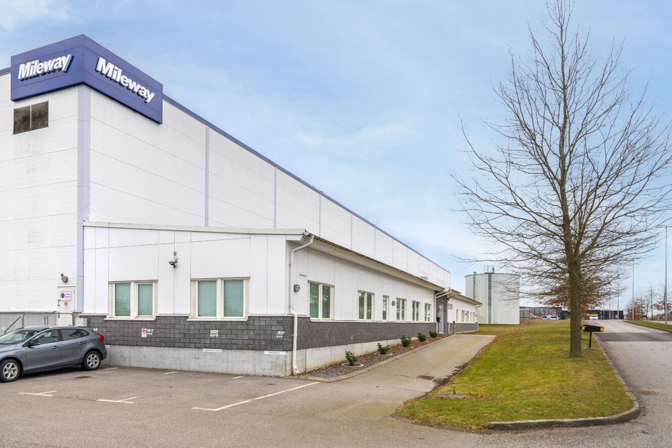 En stor vit industribyggnad med en Mileway-skylt, intill en parkeringsplats med en parkerad bil, en trottoar och ett lövfritt träd en klar dag.