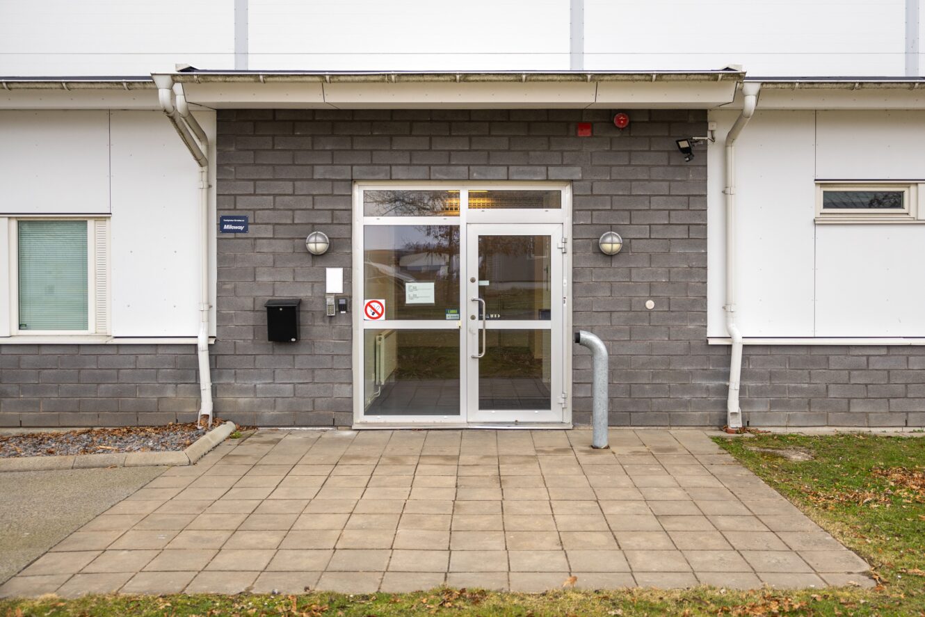 En dubbeldörr i glas till en grå tegelbyggnad med en liten markis, en brevlåda och en skylt om rökförbud på dörren. Betongplattor leder upp till entrén.