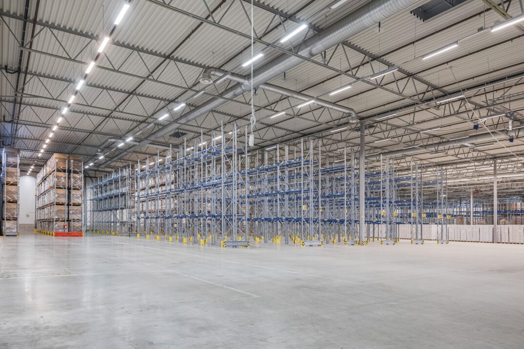 Stor, tom lagerlokal med höga metallhyllor, stark takbelysning och polerade betonggolv. På några av hyllorna till vänster står staplade lådor.