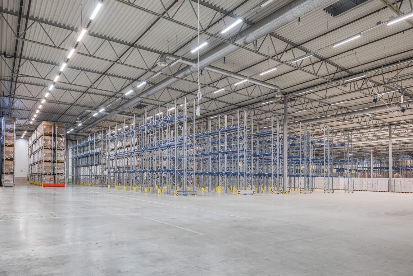 Stor, tom lagerlokal med höga metallhyllor, stark takbelysning och polerade betonggolv. På några av hyllorna till vänster står staplade lådor.