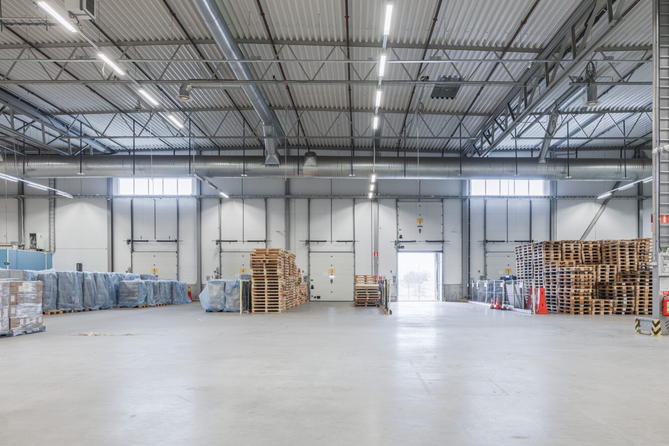 Rymlig lagerlokal med staplar av träpallar och inplastade varor längs väggarna, under högt i tak med industriell belysning.
