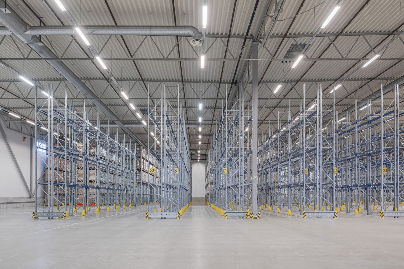 Stor, tom lagerlokal med höga förvaringsställ i metall och starka taklampor som visar upp ett organiserat industriellt förvaringsutrymme.