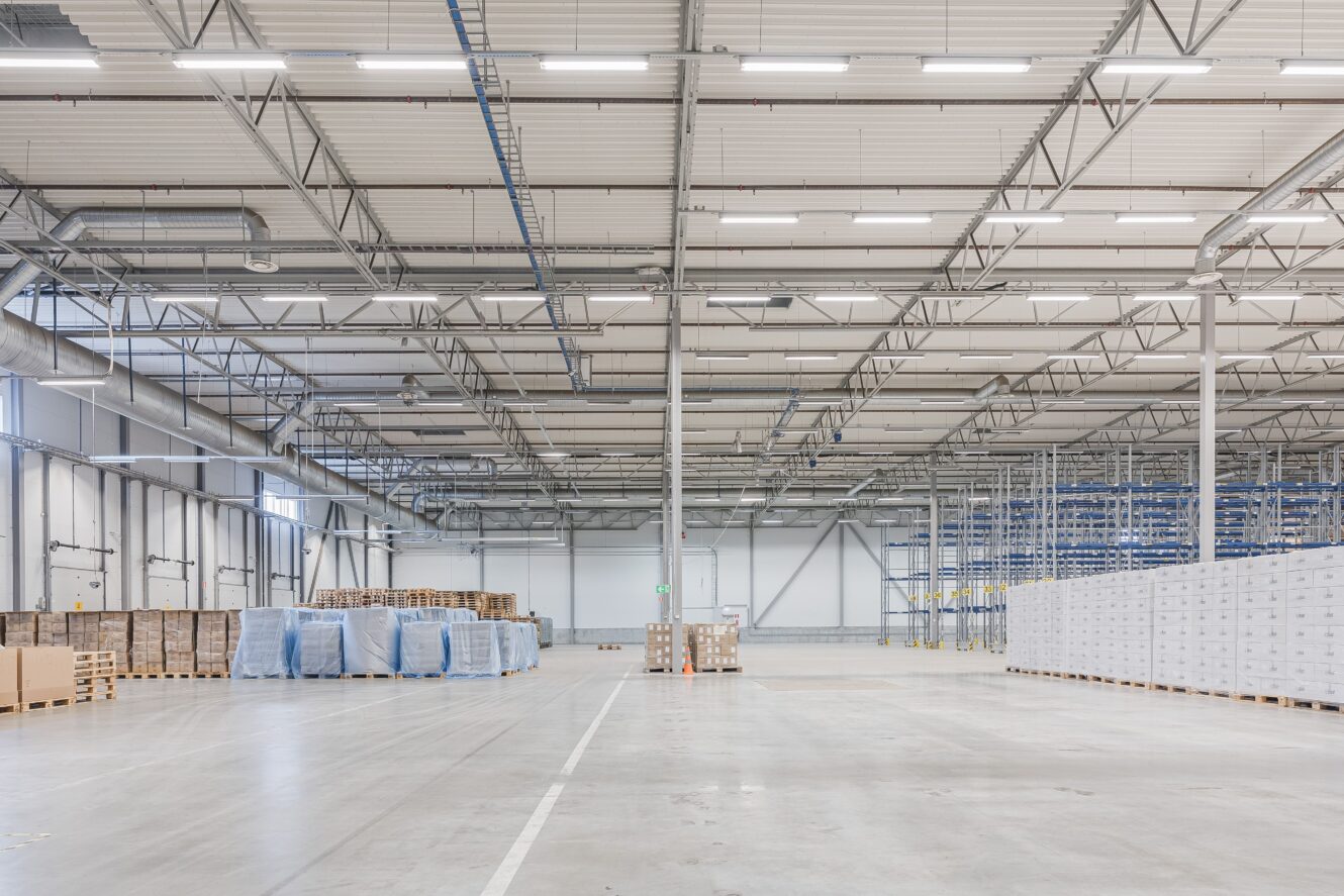 Rymlig, väl upplyst lagerlokal med staplade pallar, inslagna varor och tom golvyta under ett högt tak med synliga bjälkar.