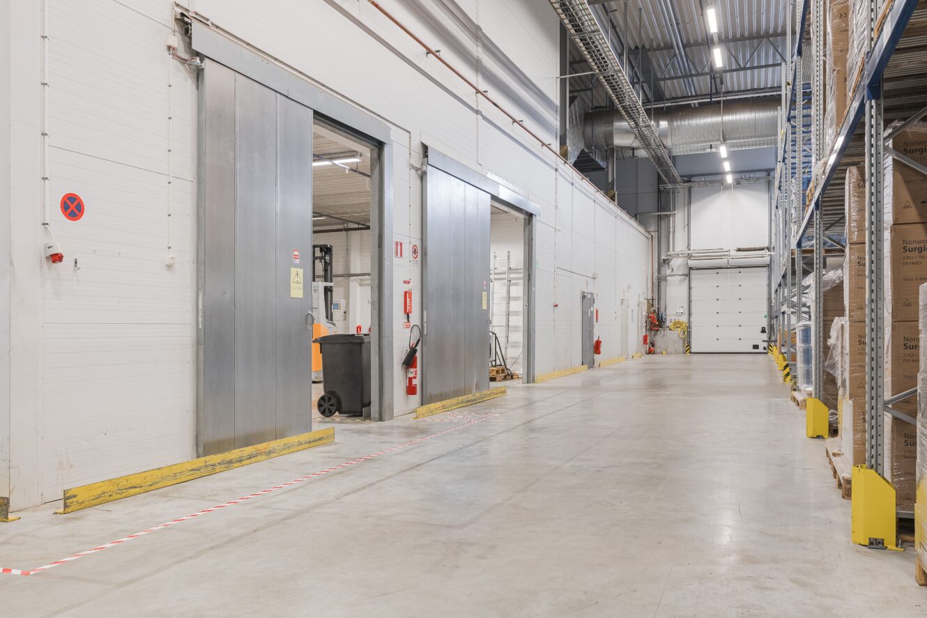 En rymlig, ren lagerlokal med stora skjutdörrar i metall, staplade lådor på ställningar och ett betonggolv.