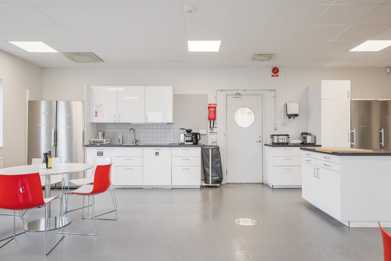 Modernt fikarumskök med vita skåp, rostfria vitvaror, röda stolar runt ett bord och olika köksmaskiner på bänkskivorna. Golvet är grått och utrymmet är väl upplyst.