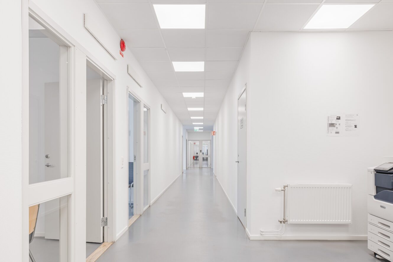 En lång, ljus korridor i en modern byggnad med vita väggar, flera dörrar, en väggradiator och en kopiator till höger.