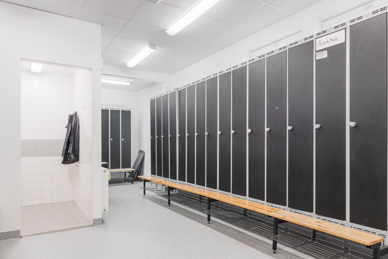 Ett rent och modernt omklädningsrum med svarta skåp, träbänkar och ett kaklat duschutrymme syns till vänster.
