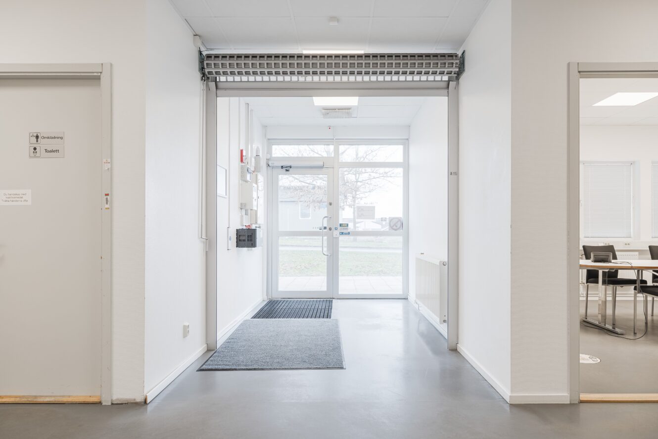 En ren, vit hall med en grå golvmatta leder till en ingång med glasdörr; intilliggande rum på vardera sidan innehåller kontorsmöbler.