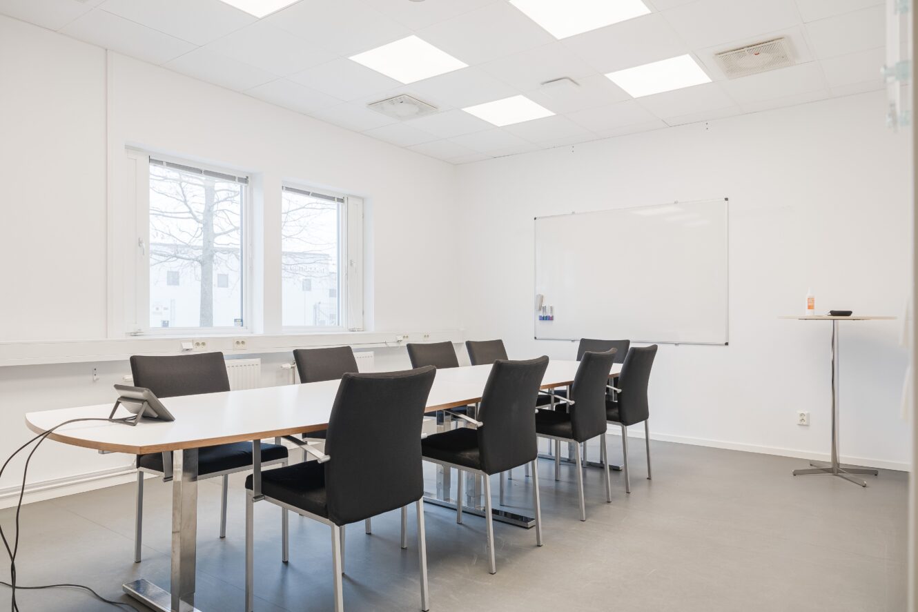 Ett modernt konferensrum med ett långt vitt bord, åtta svarta stolar, två fönster, en whiteboard på väggen och ett litet runt bord i hörnet.