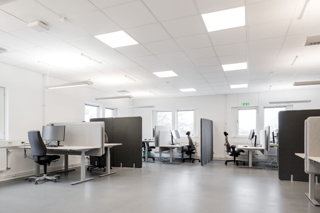 Modernt kontorslandskap med flera bås, skrivbord, stolar och datorskärmar. Utrymmet är tomt och väl upplyst med stora fönster och taklampor.
