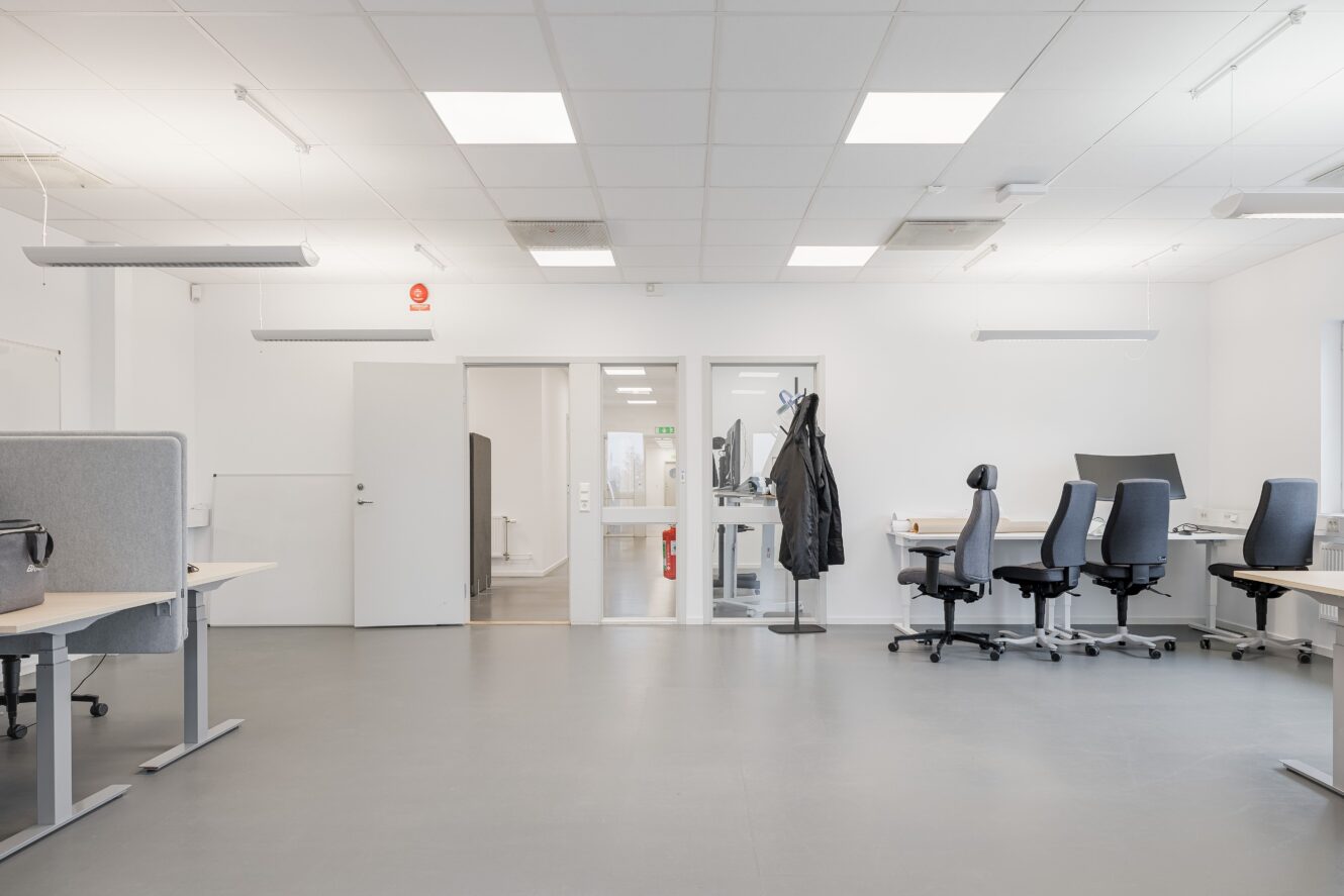 Ett modernt, tomt kontor med flera skrivbord och kontorsstolar, vita väggar, stark belysning och en kappa hängande vid dörren.