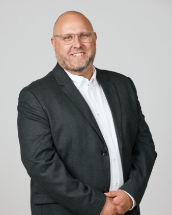 En skallig man med glasögon och skägg, klädd i mörk kostym och vit skjorta, står mot en ljus bakgrund och ler mot kameran.