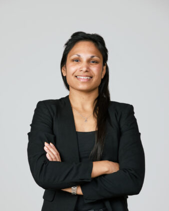 En kvinna med långt mörkt hår och svart kavaj står med armarna i kors och ler mot kameran mot en ljus bakgrund.