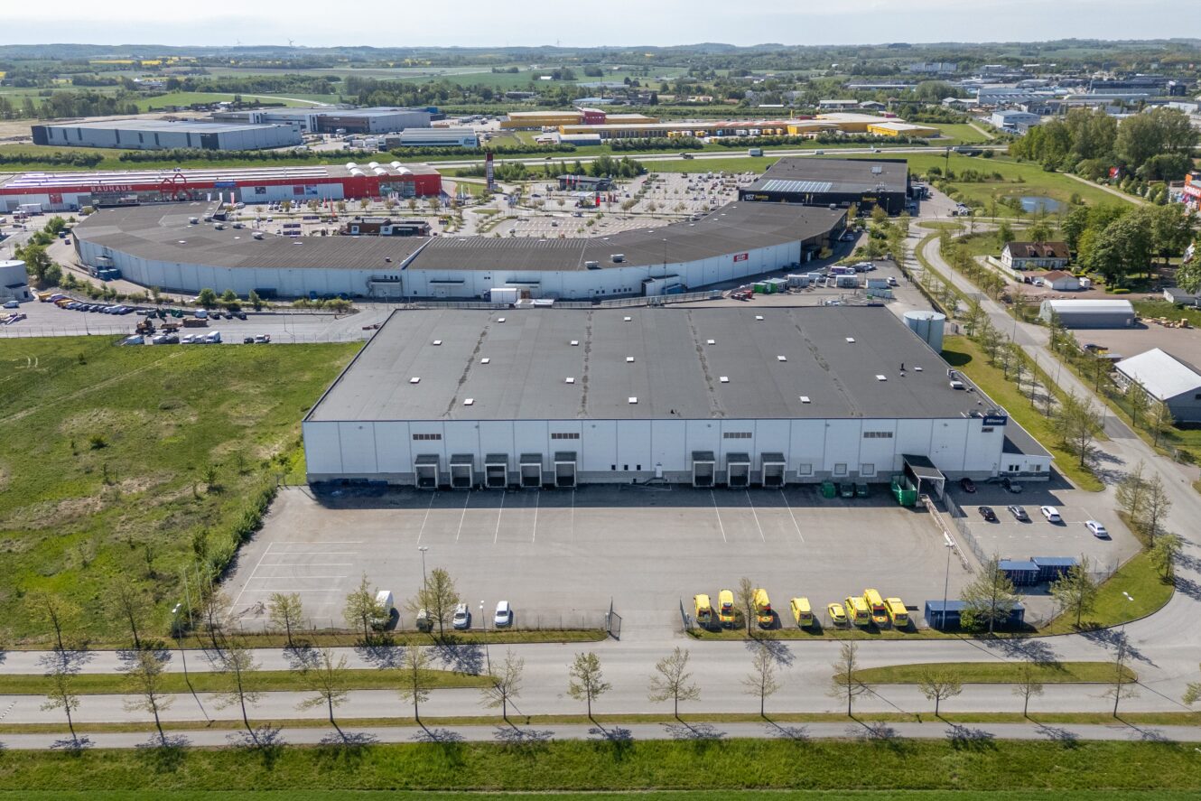 Flygfoto över en stor lagerlokal med lastkajer och parkerade lastbilar i ett industriområde, omgiven av andra kommersiella byggnader och grönska.
