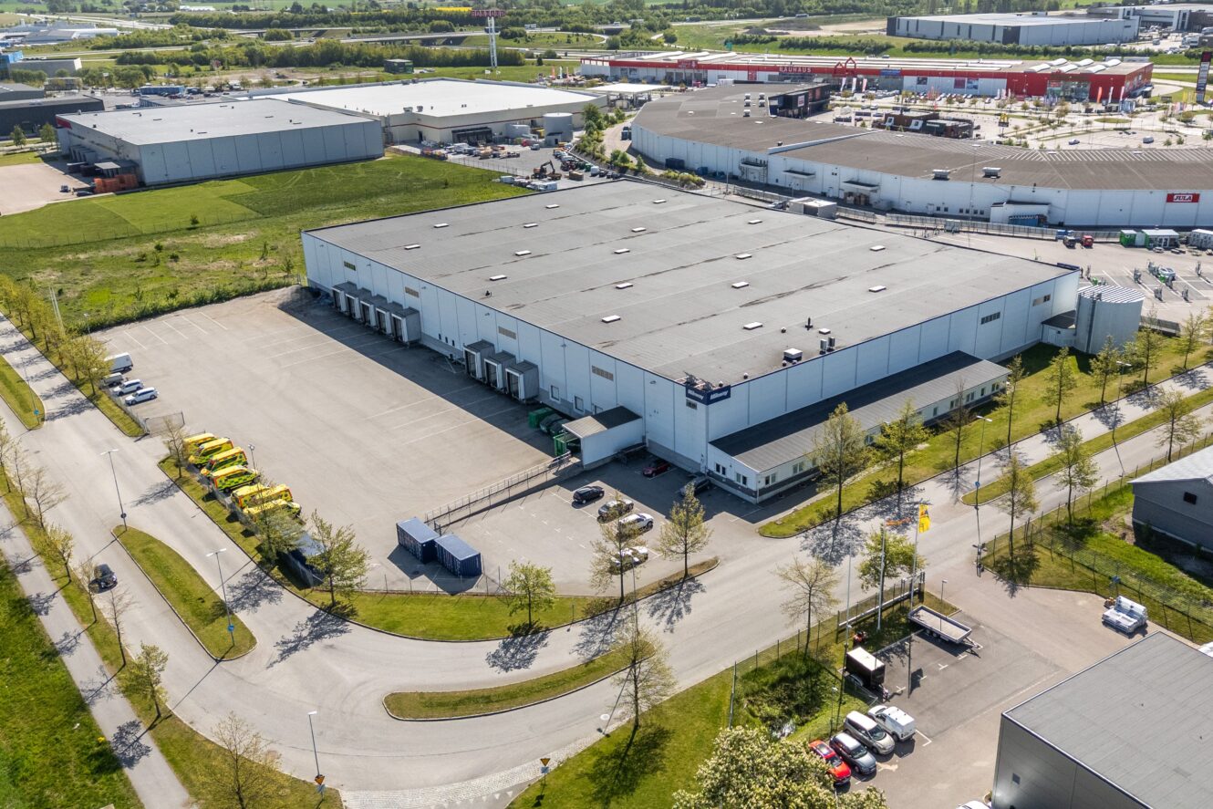 Flygfoto över en stor vit lagerlokal med lastkajer, lastbilar parkerade i närheten samt omgivande vägar och grönområden i en industripark.