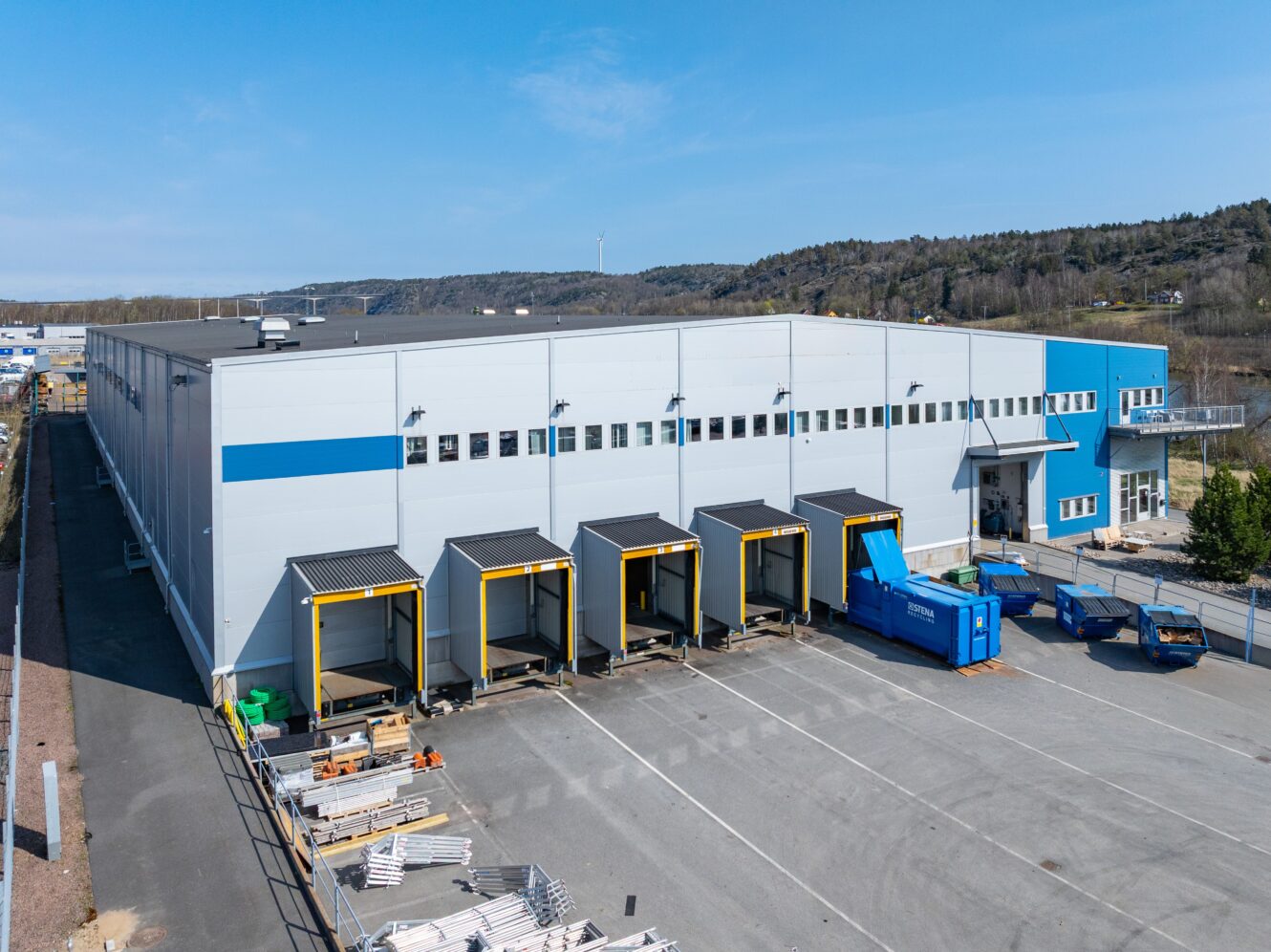 Stort industrilager med fyra lastbryggor, en blå återvinningscontainer och byggmaterial på parkeringen, mot en bakgrund av träd och kullar.