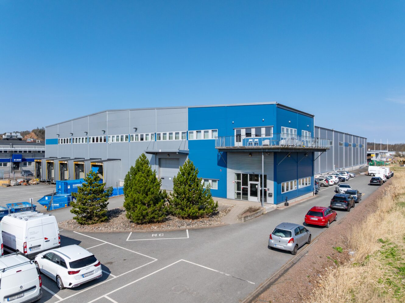 Stort blått och grått industrilager med lastkajer och parkerade bilar på den intilliggande tomten under en klar himmel.