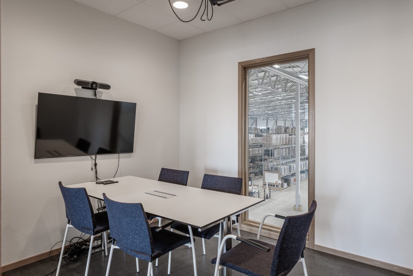 Ett litet konferensrum med ett bord, fem stolar, en väggmonterad TV och ett fönster med utsikt över en lagerlokal med hyllor och utrustning.