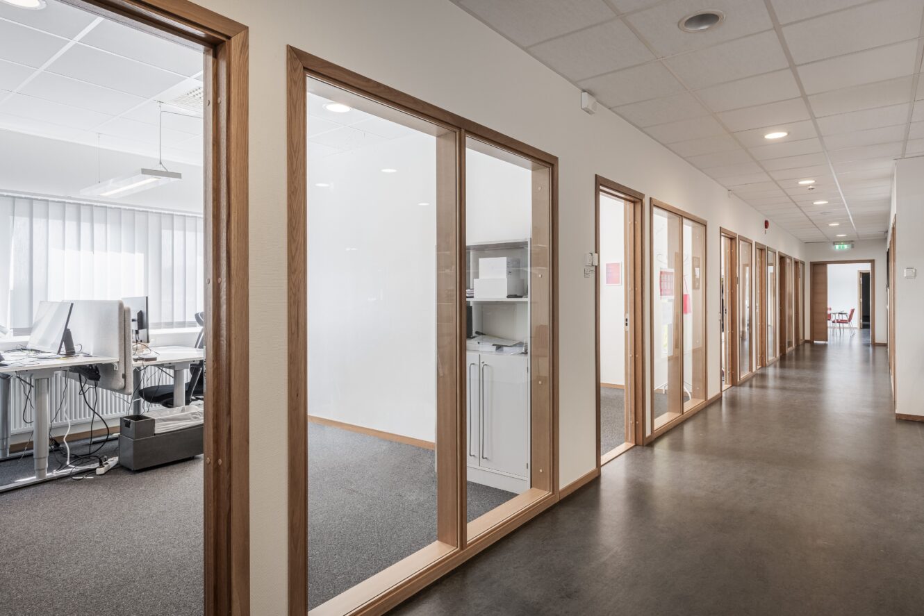 En modern kontorshall med glasväggar, synliga skrivbord, datorer och kontorsutrustning; stark belysning och minimal inredning.