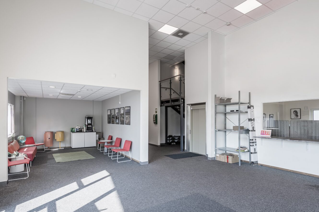 Rymligt rum med röda stolar, kaffestation, hyllor och en trappa som leder till ett övre plan; vita väggar och heltäckningsmatta på golvet.