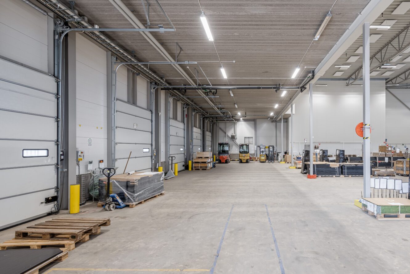 Rymlig lagerlokal med pallar, staplade varor, gaffeltruckar och utrustning längs väggarna under starka taklampor.